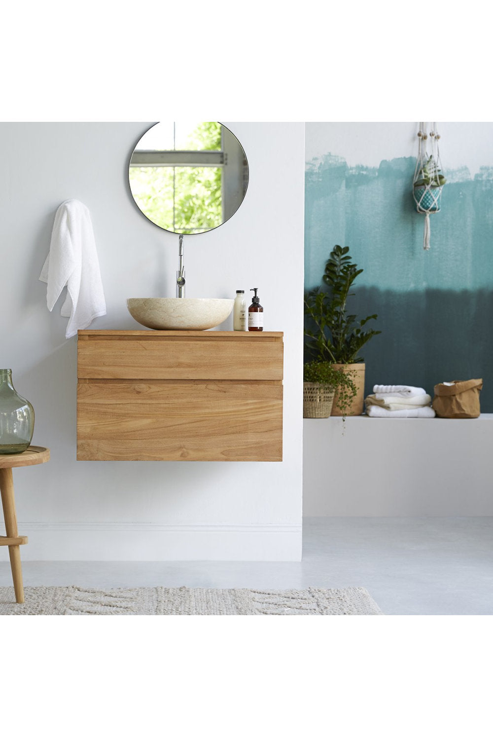 Modern floating teak single-sink vanity with natural wood finish and cream stone marble top, fitted with a round vessel sink and chrome wall faucet; styled with a round mirror, white towel and potted plants.
