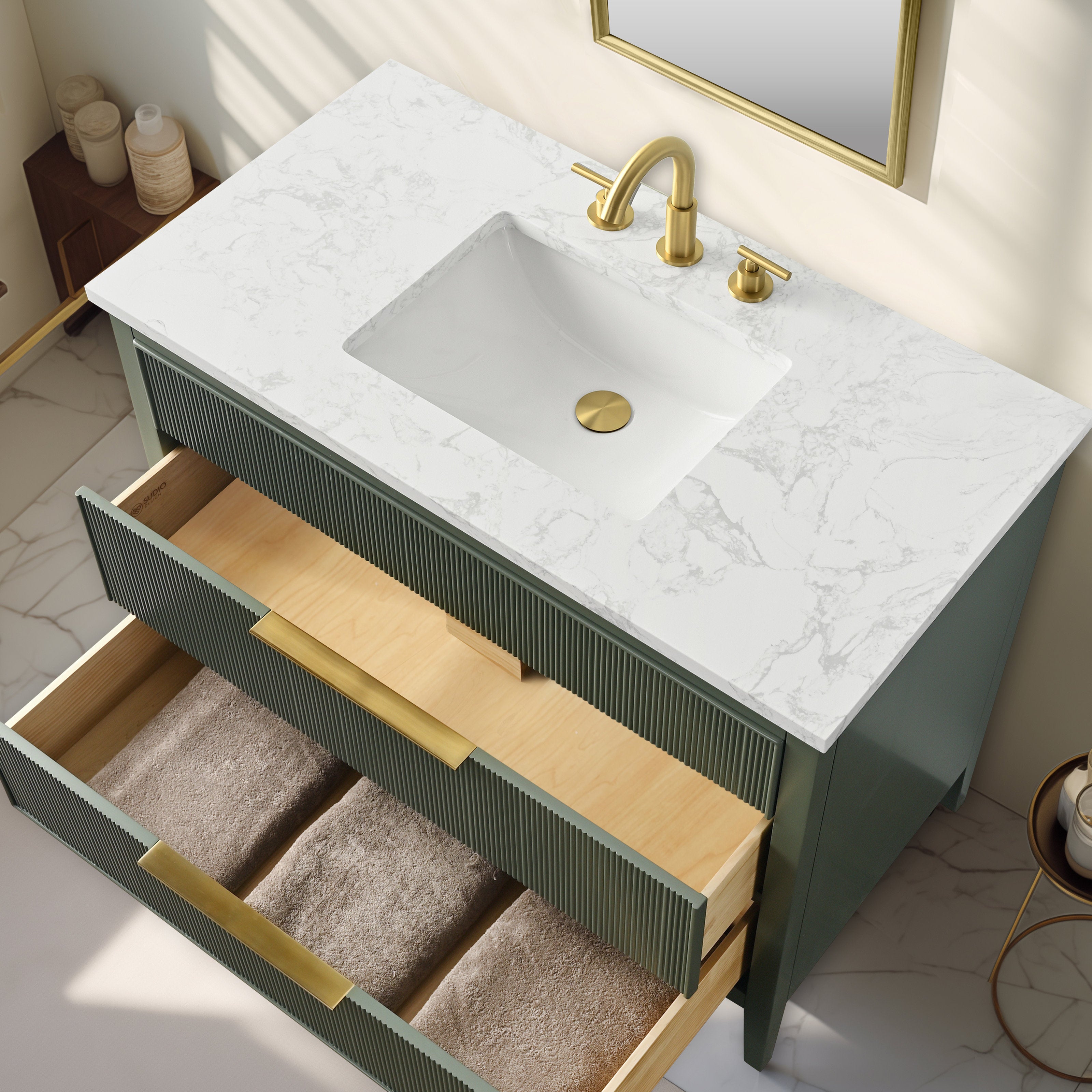 Top-down view of a modern solid wood single-sink bathroom vanity with a white stone countertop and undermount ceramic sink, green fluted drawer fronts, brass faucet and handles, open drawers showing wood interiors and folded towels.