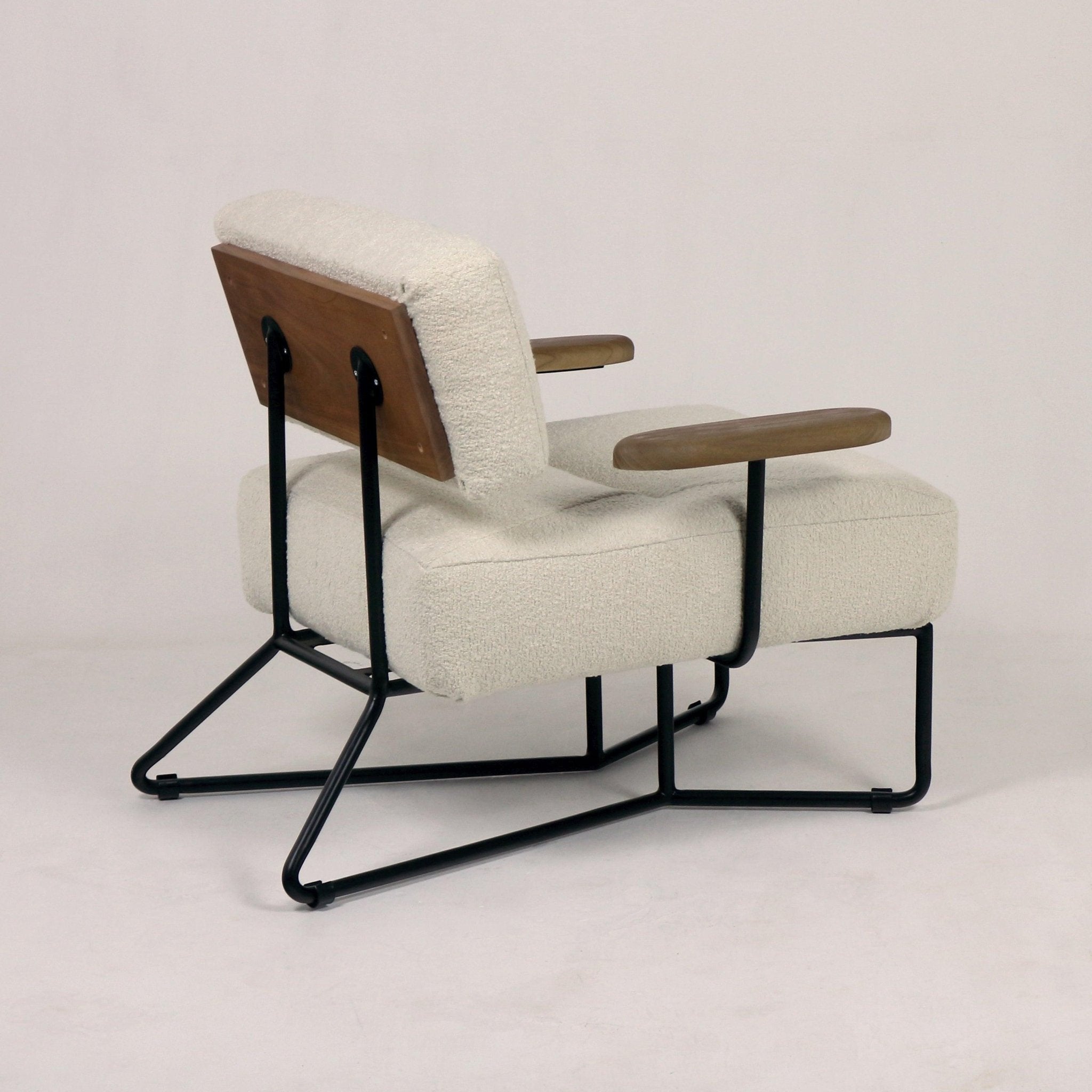 Side view of a mid-century modern low lounge chair upholstered in beige boucle fabric with padded seat and back, wooden back panel and sculpted wooden armrests, supported by a black metal sled-style frame.
