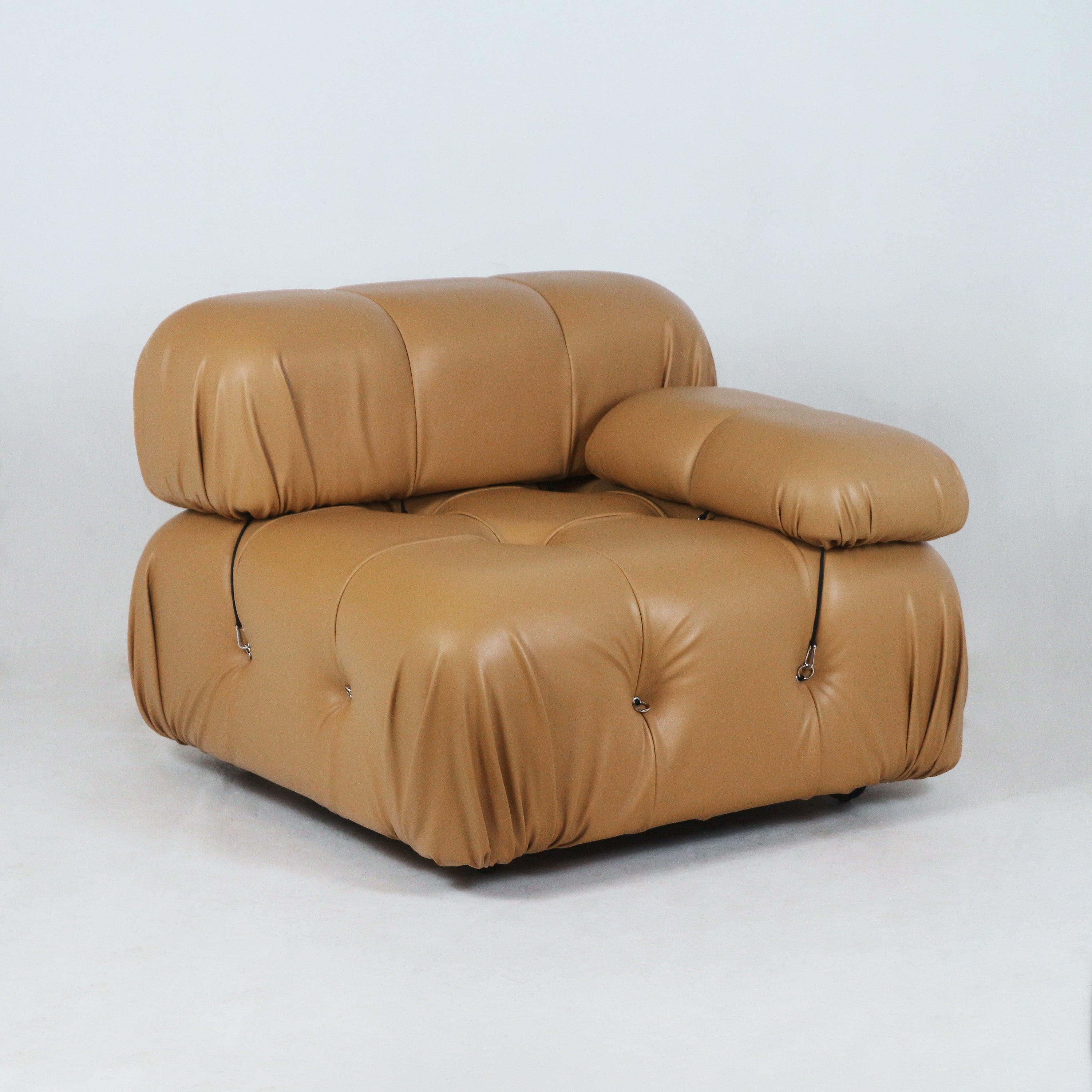 Low-profile tan leather modular sofa section with rounded, tufted cushions, a single padded armrest and visible black straps with metal ring details against a neutral studio background.
