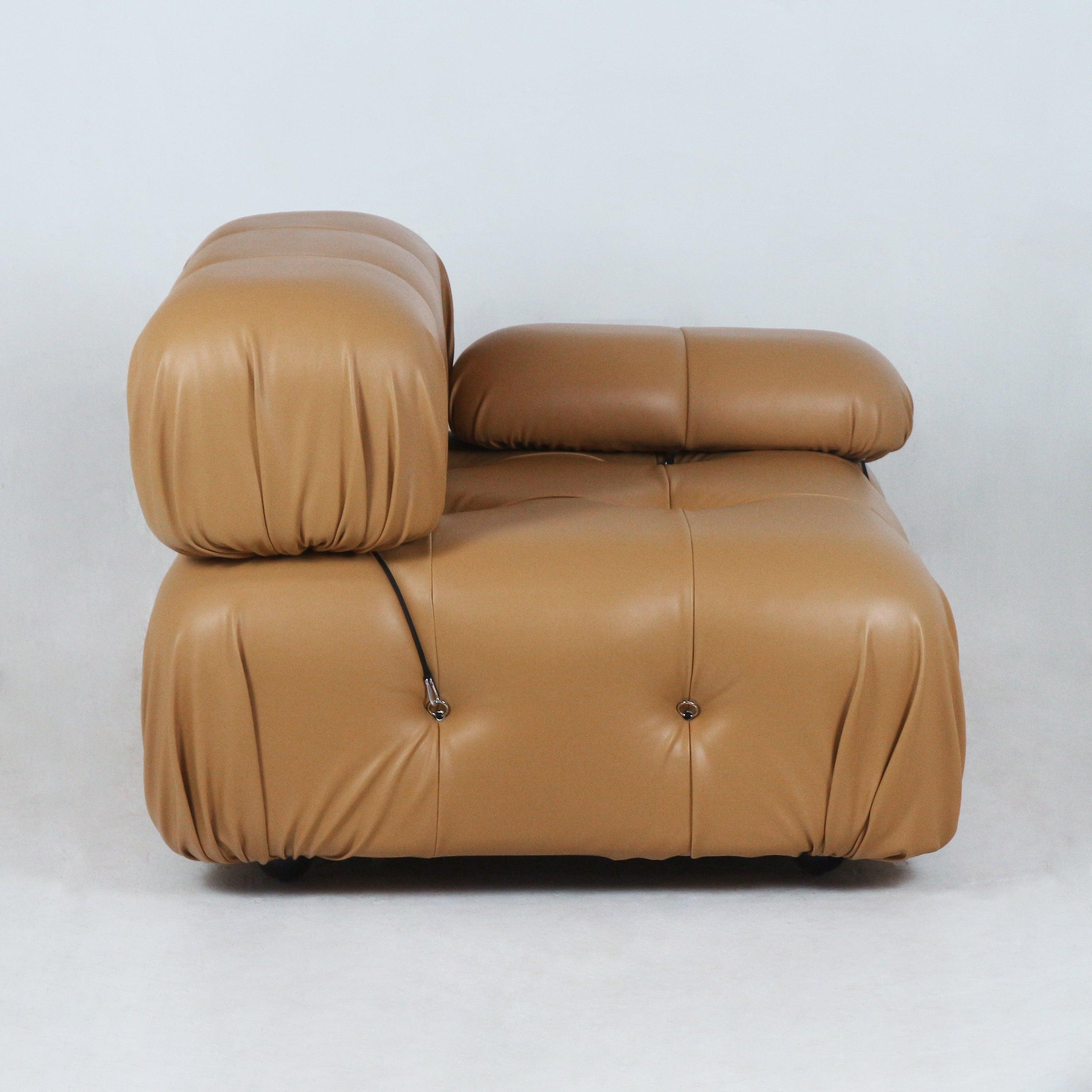 Low-profile modern tan leather modular sofa section with rounded tufted cushions, adjustable headrest and visible black strap with metal hardware on a white studio background.