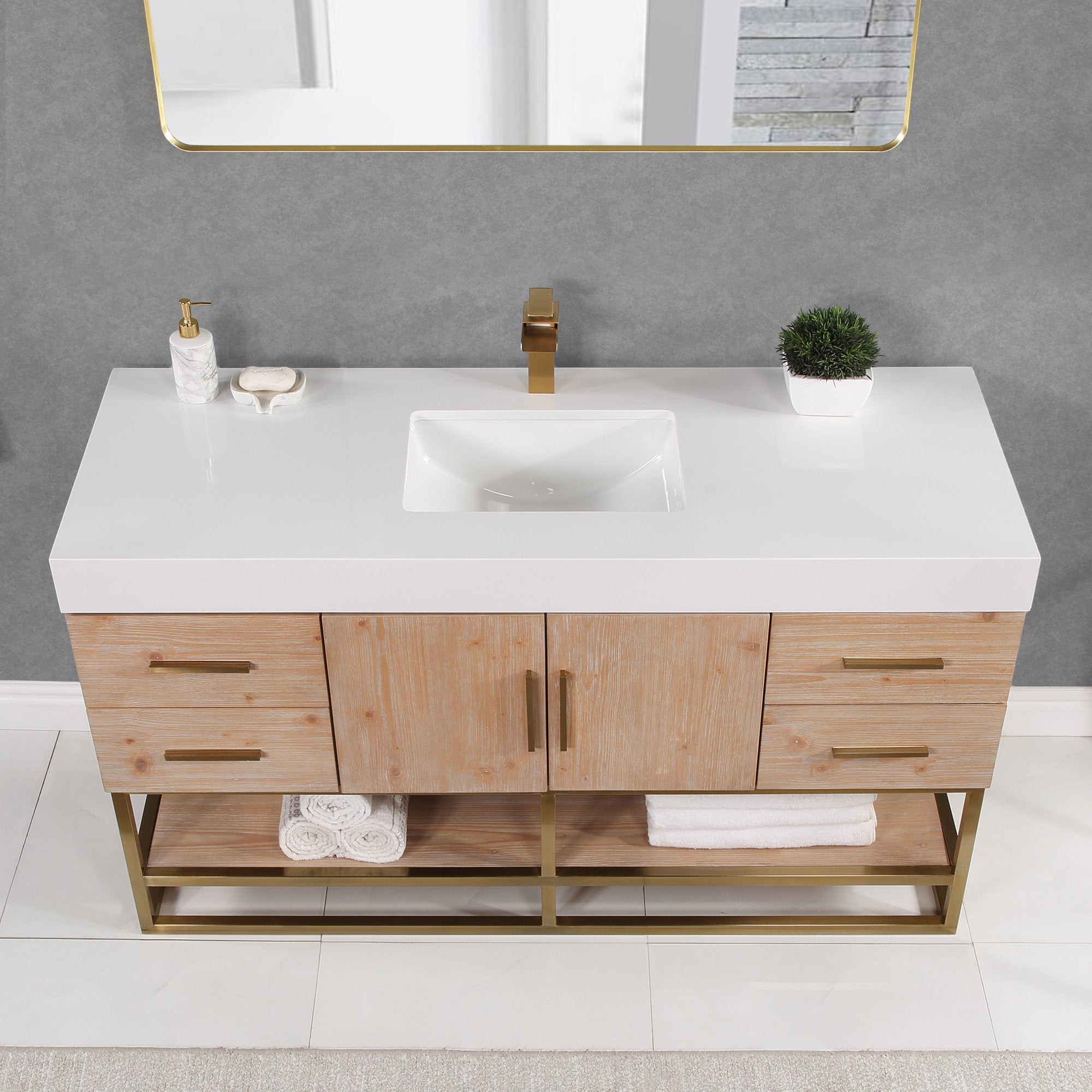 Modern single-sink bathroom vanity with a glossy white composite stone top and rectangular undermount sink, light natural wood cabinet, brass-finish faucet and handles, gold metal base shelf with folded towels and a small potted plant.