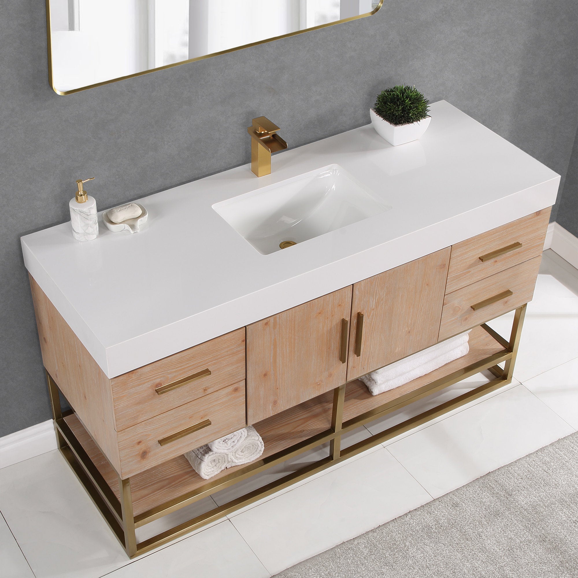 Modern 60-inch single bathroom vanity with white composite stone top and rectangular undermount sink, light natural wood cabinet, brass-gold faucet and hardware, open brass metal base shelf with rolled white towels, soap dispenser and small plant.