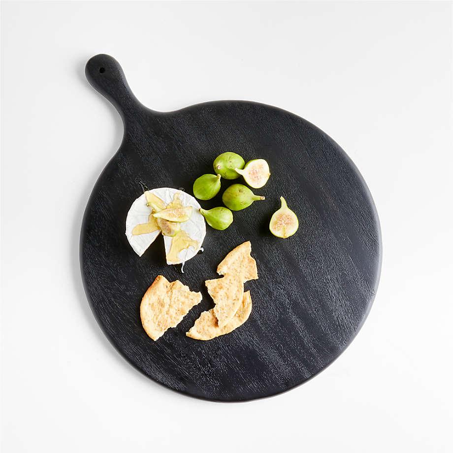 Round black ash wood paddle cutting board photographed from above holding a wheel of brie with honey, halved green figs and broken crackers on a white background.