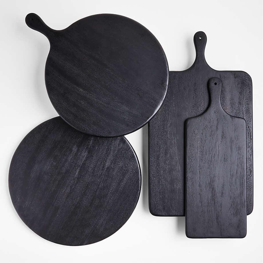 Overhead view of four black teak serving boards — two round paddle-style boards and two rectangular paddle boards — showing wood grain and handle holes, arranged on a white background for a contemporary kitchen display.