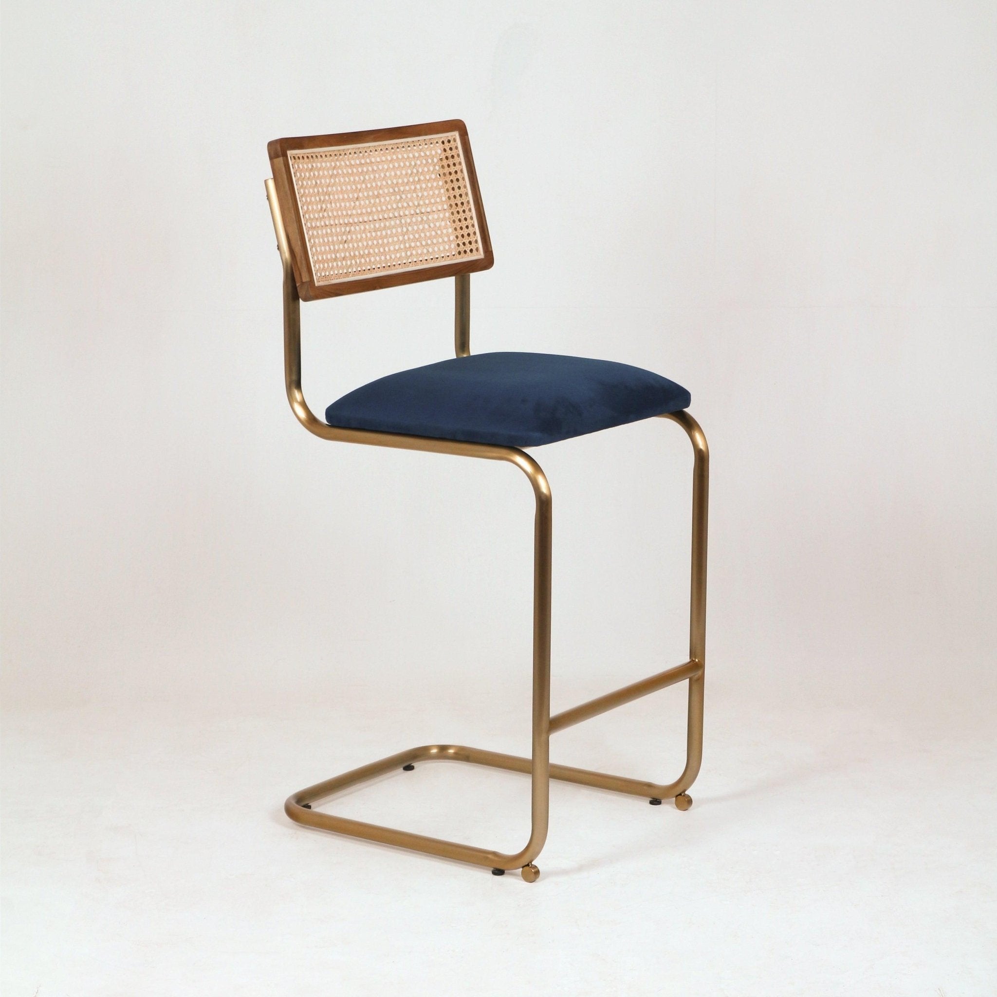 Cantilever counter-height bar stool with a brass-finish metal frame, navy velvet upholstered seat and natural cane-backed wooden backrest shown on a white studio background.