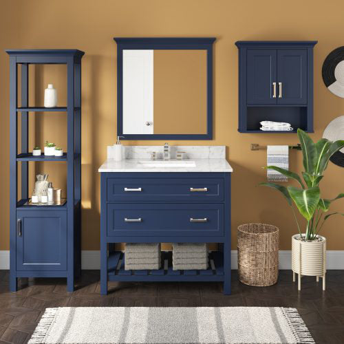 Navy blue solid wood single-sink bathroom vanity with white marble countertop, matching framed mirror, tall open shelving unit and wall cabinet, chrome hardware, woven basket and potted plant against a warm tan wall.
