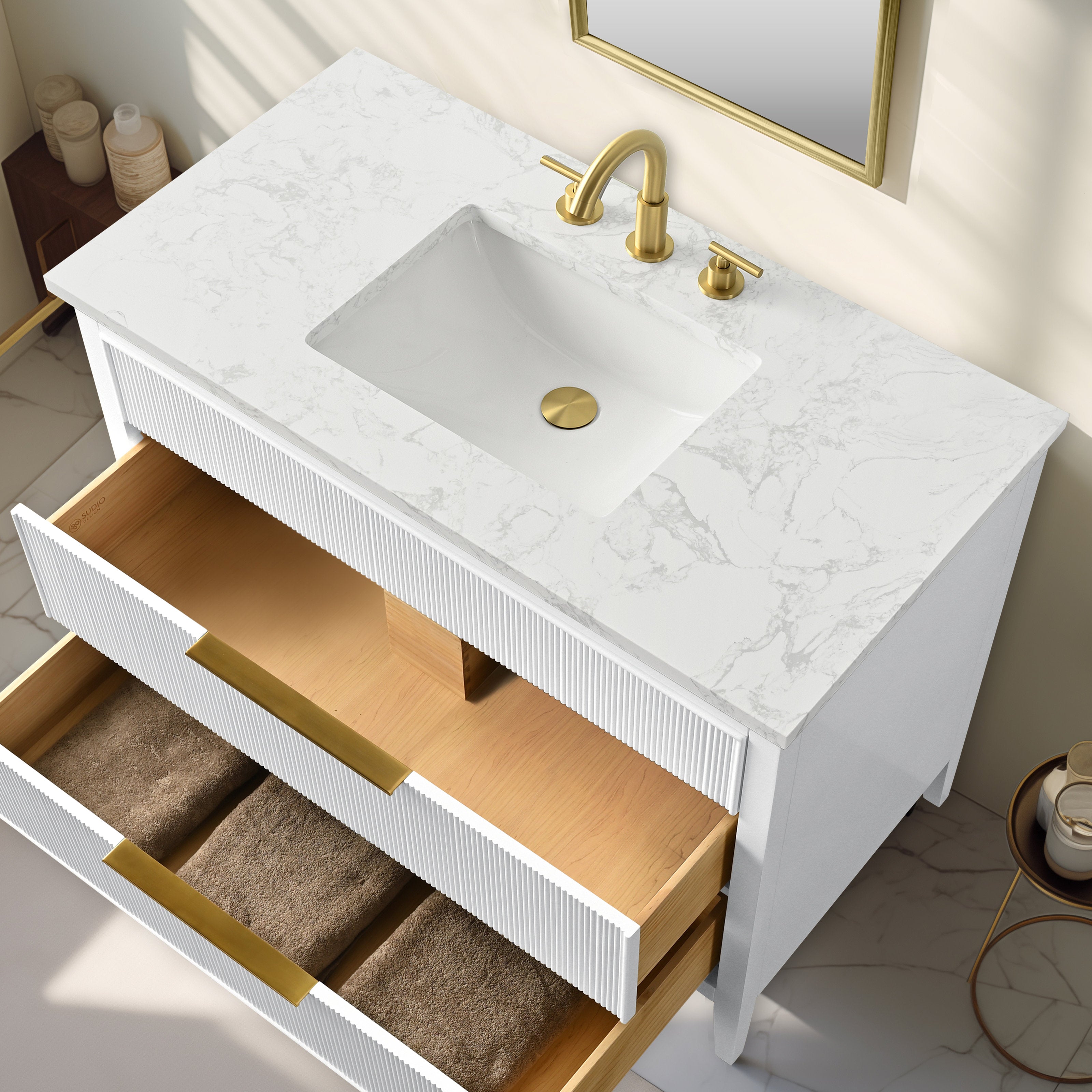 Overhead view of a modern solid wood bathroom vanity with a white stone-look countertop, rectangular ceramic sink, brass-finish faucet and drain, fluted white drawer fronts with brass handles and open drawers showing wood interior and brown towels.