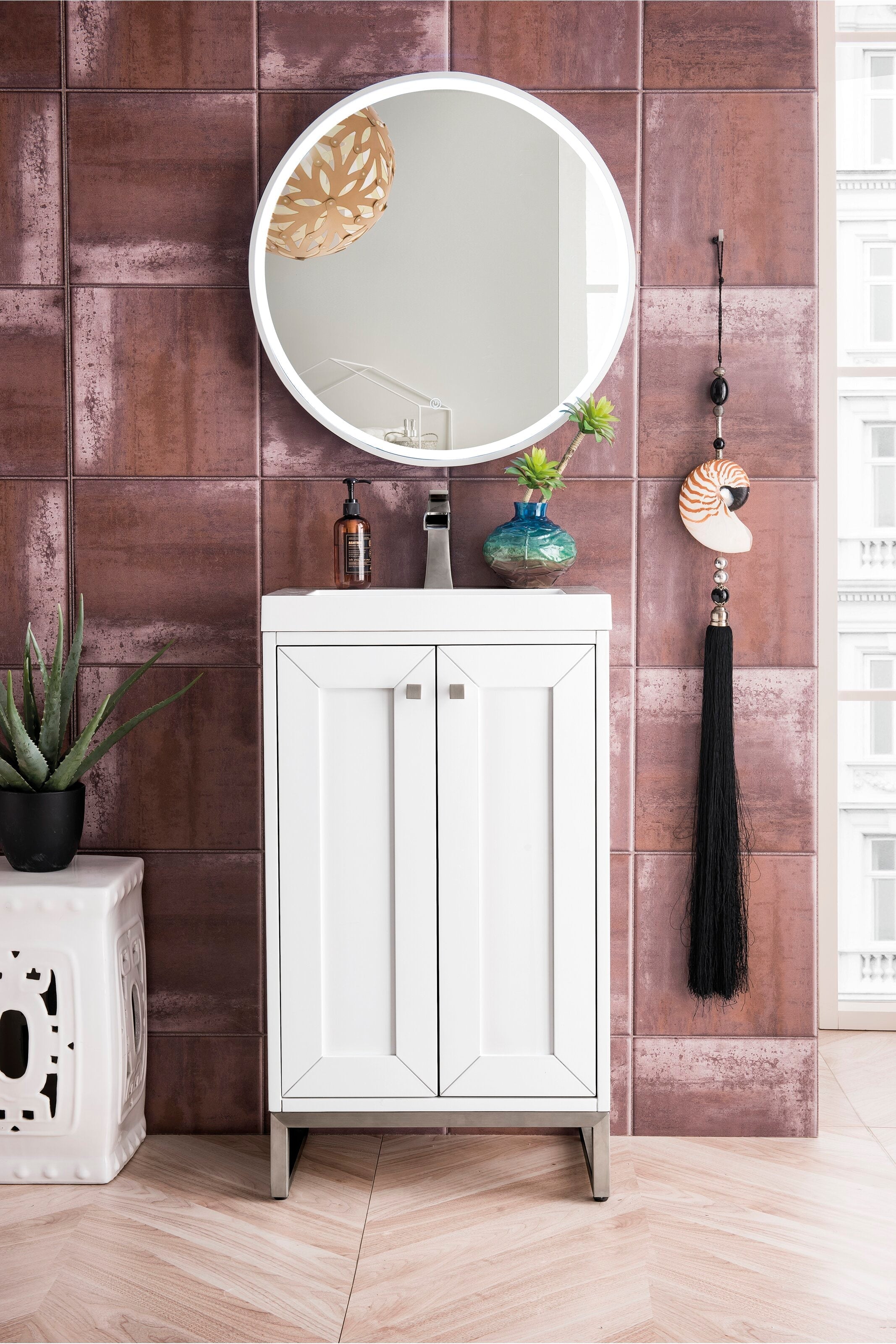 Freestanding modern white solid wood bathroom vanity with stone top and single ceramic sink, brushed-nickel faucet, round illuminated mirror, amber soap dispenser and small succulent vase against textured stone-colored tile wall.