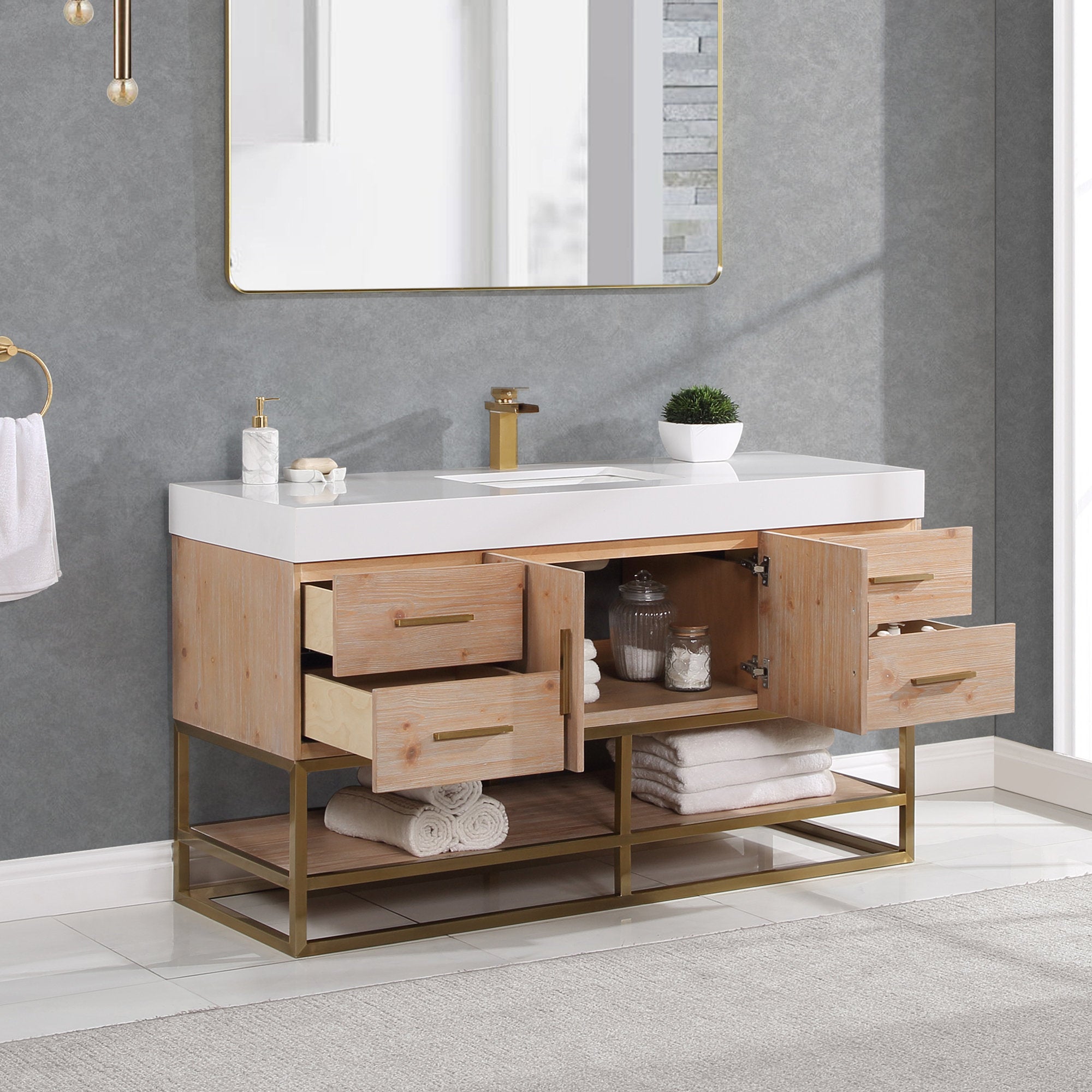 Modern 60-inch single-sink bathroom vanity with a white composite stone top and rectangular undermount sink, natural wood cabinet with brass hardware, open drawers and gold metal base shelf holding towels and jars.