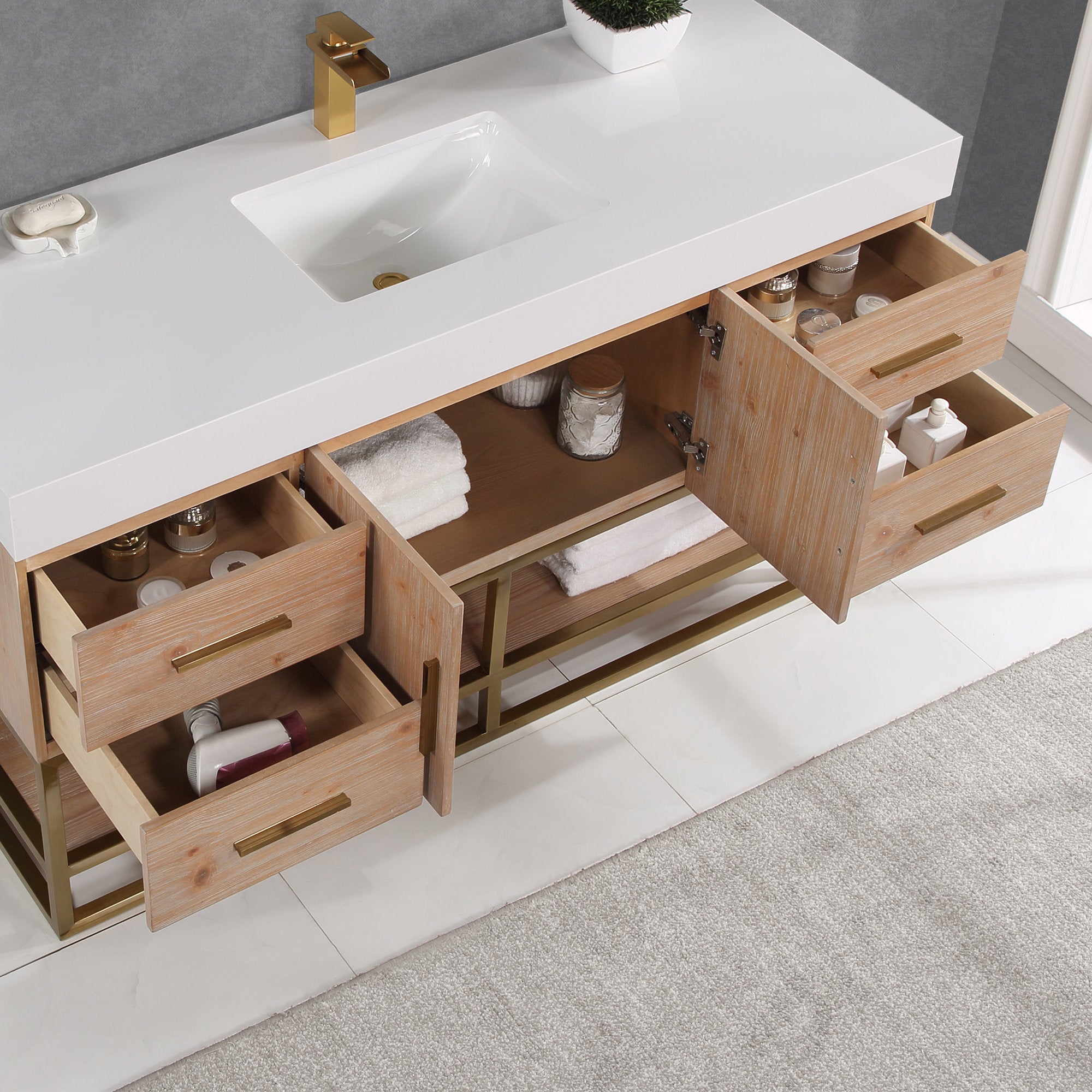 Overhead view of a modern 60-inch single-sink bathroom vanity with a white composite stone top and rectangular undermount sink, light natural wood cabinet on a gold metal base, brass faucet and handles, open drawers with towels and toiletries.