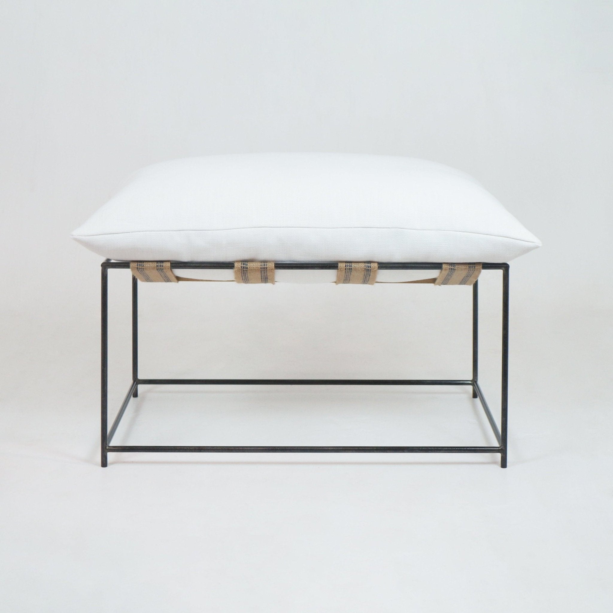 Square ottoman with a plush white linen cushion atop a slim dark metal rectangular frame, secured by exposed natural woven straps, photographed on a plain white background.